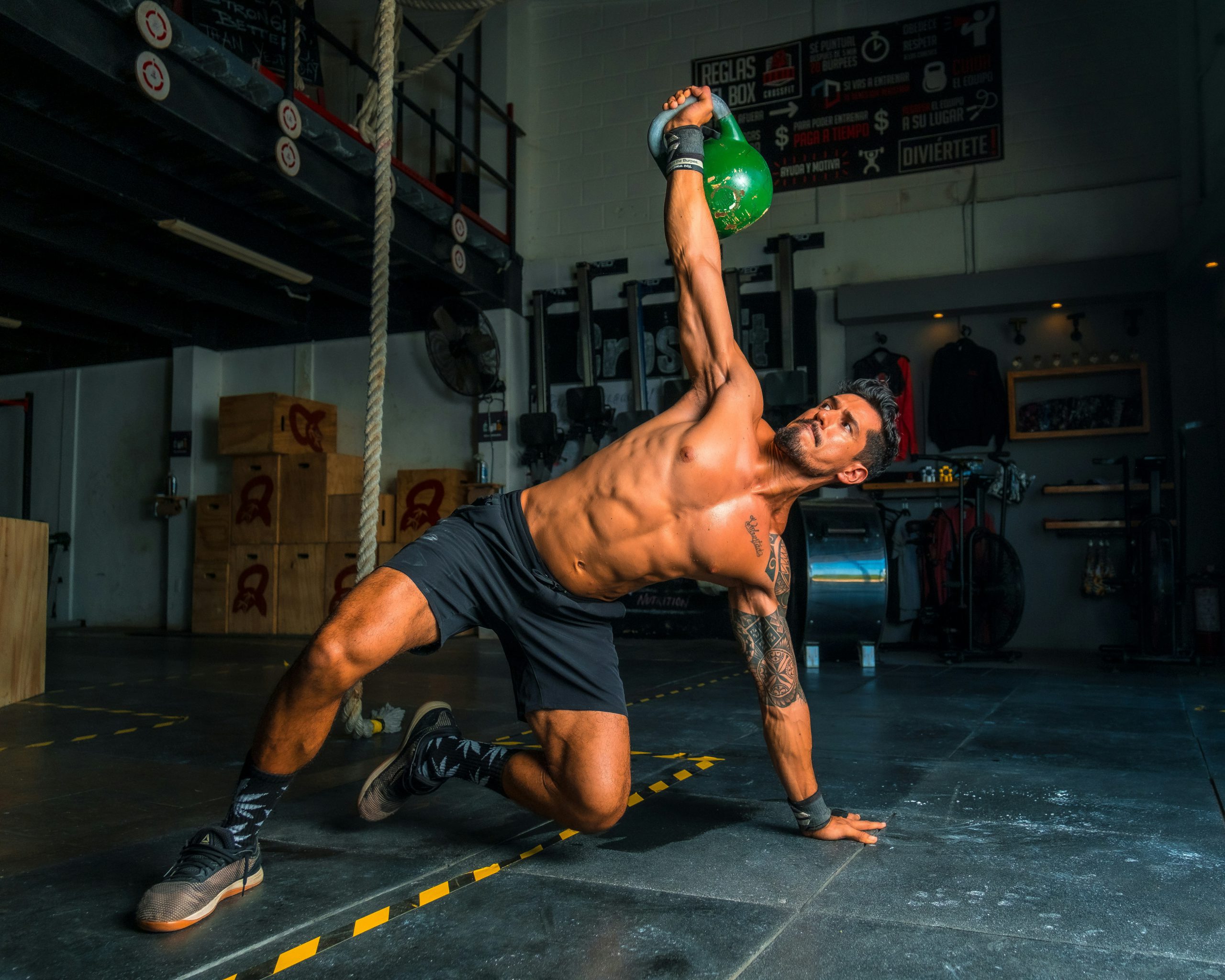 Uomo a torso nudo mentre esegue un esercizio con kettlebell in palestra, in posizione bassa e con un braccio sollevato.