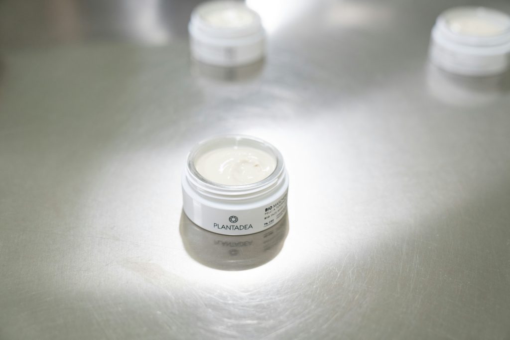 Un barattolo aperto di crema cosmetica è posizionato su una superficie metallica, con altri contenitori sfocati sullo sfondo