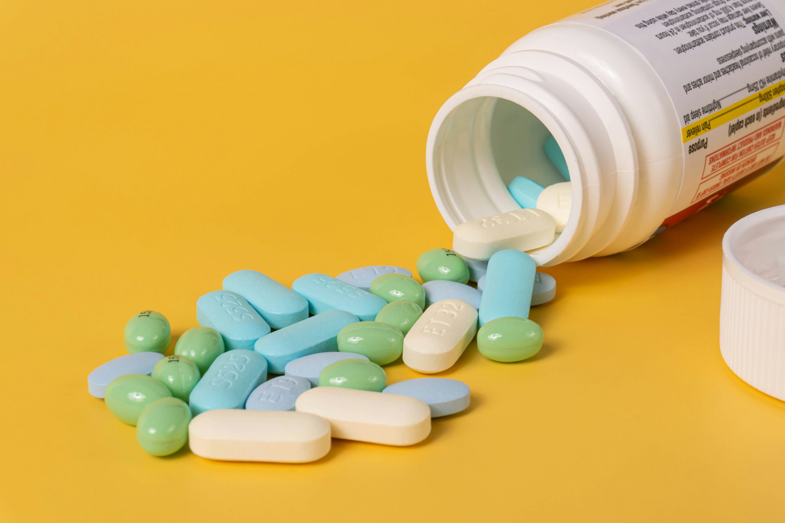 Un flacone di medicinali aperto da cui fuoriescono diverse compresse e pillole nei colori blu, verdi e bianchi su uno sfondo giallo