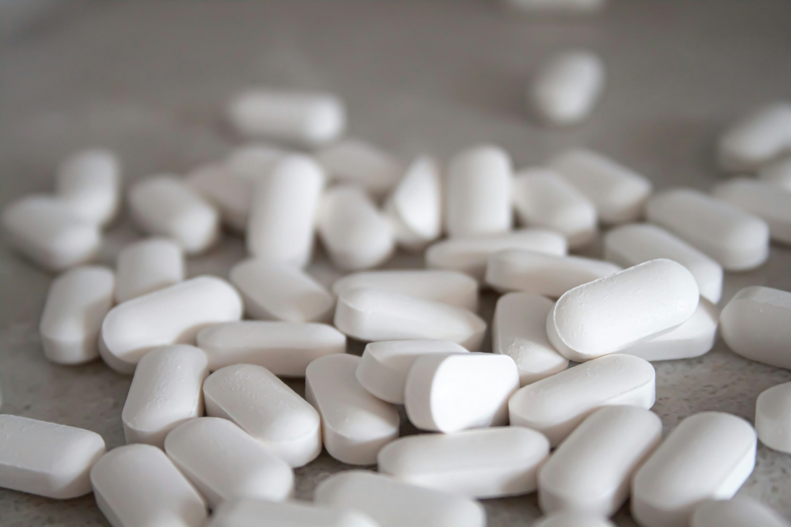 Numerose compresse bianche di forma ovale sparse su una superficie chiara, rappresentazione di farmaci o integratori.