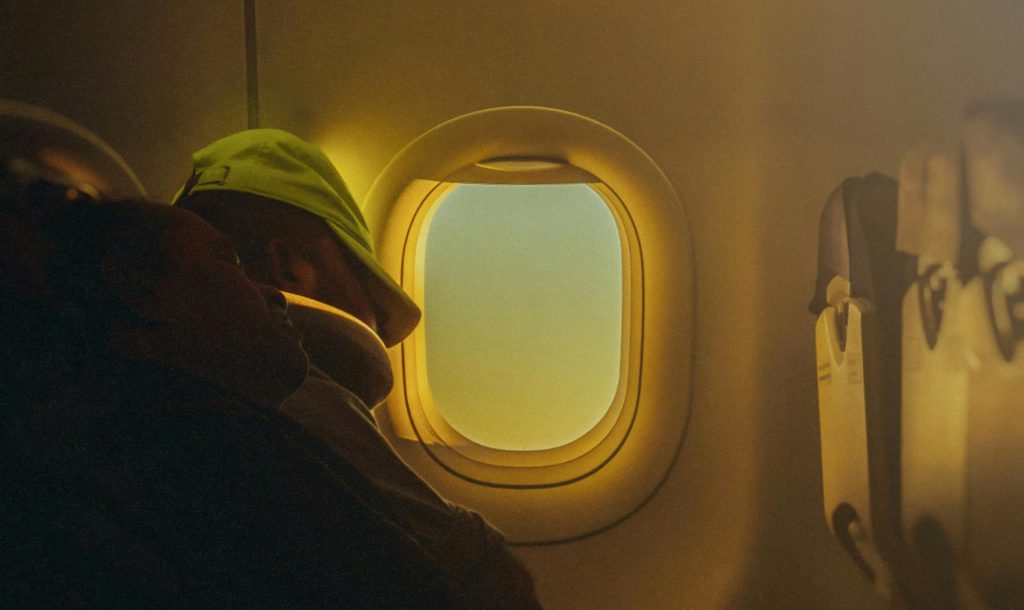 Passeggero che dorme in aereo vicino al finestrino illuminato dal sole, a simboleggiare la gestione del jet lag e dei ritmi circadiani nei voli transoceanici.