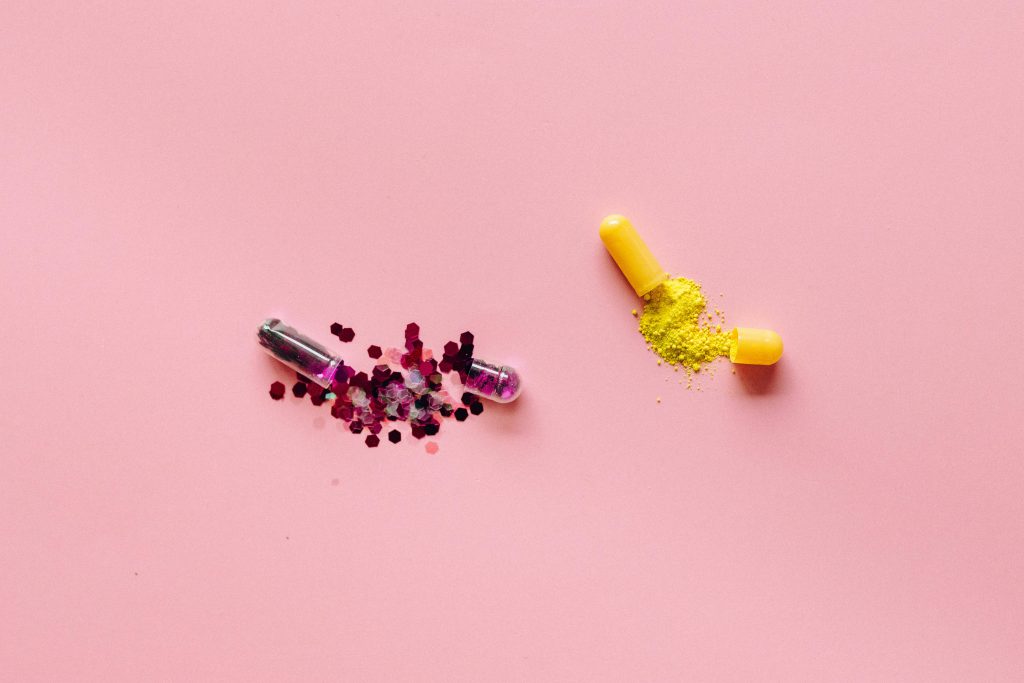 Due capsule aperte su fondo rosa che rilasciano polvere gialla e glitter, a simboleggiare l'integrazione di peptidi e il biohacking a livello cellulare.